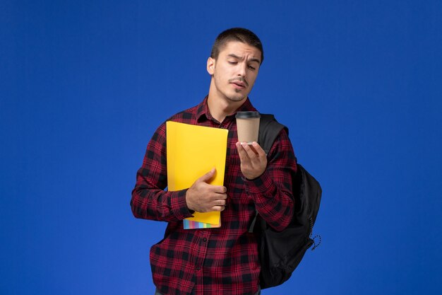 Vooraanzicht van mannelijke student in rood geruit overhemd met rugzak met gele bestanden en koffie op de blauwe muur
