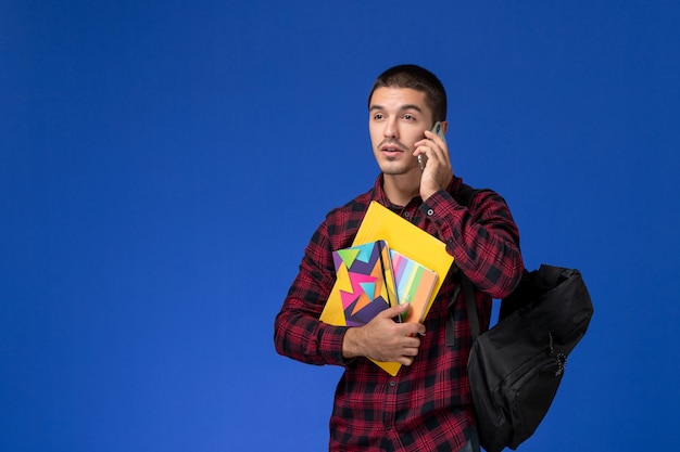 Vooraanzicht van mannelijke student in rood geruit overhemd met voorbeeldenboek van de rugzakholding en dossiers die aan de telefoon op blauwe muur spreken