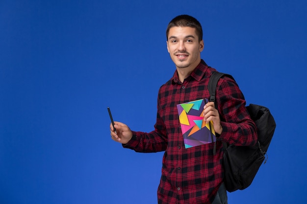 Vooraanzicht van mannelijke student in rood geruit overhemd met voorbeeldenboek van de rugzakholding en pen op lichtblauwe muur