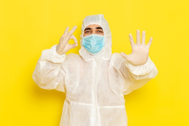Gratis foto vooraanzicht van mannelijke wetenschappelijke werker in speciaal beschermend pak en met masker dat zijn palm op lichtgele muur toont