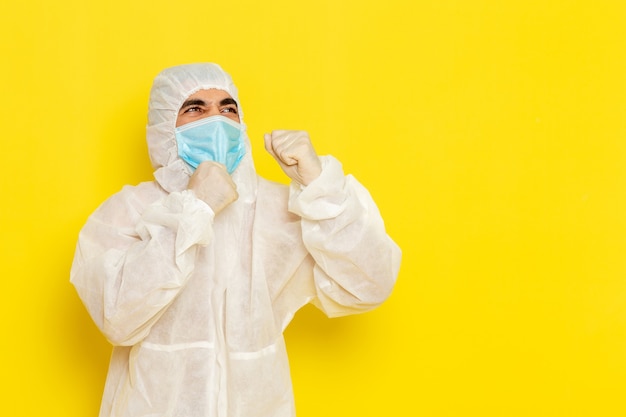 Vooraanzicht van mannelijke wetenschappelijke werker in speciaal beschermend pak en met masker poseren op de lichtgele muur