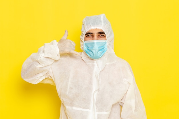 Vooraanzicht van mannelijke wetenschappelijke werknemer in speciaal beschermend wit pak en met masker poseren op de gele foto van het de chemiekleurengevaar van de bureauwetenschapper