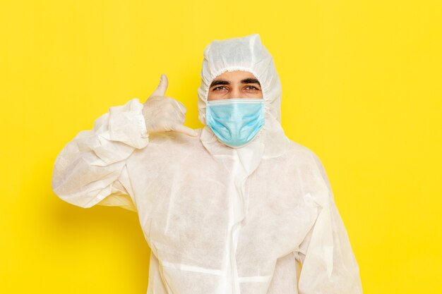 Vooraanzicht van mannelijke wetenschappelijke werknemer in speciaal beschermend wit pak en met masker poseren op de gele foto van het de chemiekleurengevaar van de bureauwetenschapper