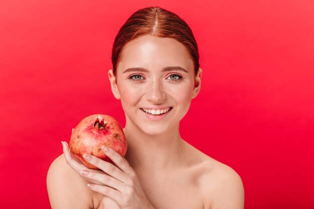 Vooraanzicht van meisje met granaat geluk uitdrukken. Studio shot van lachende gember vrouw met granaatappel geïsoleerd op rode achtergrond.