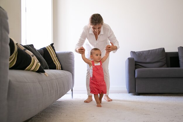 Vooraanzicht van moeder die kleine meisjeshanden houdt en helpt om te lopen. Schattig blootsvoets babymeisje in rode werkbroeken korte broek leren wandelen in de woonkamer met hulp van moeder. Familie tijd en eerste stap concept