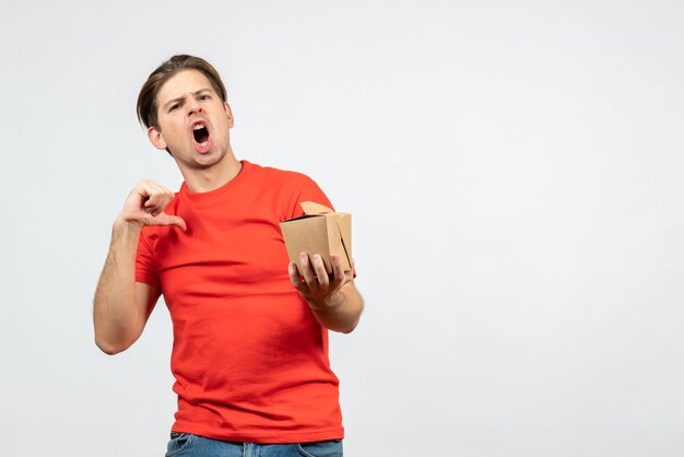 Vooraanzicht van nerveuze jonge kerel in rode blouse die kleine doos houdt die op witte achtergrond richt