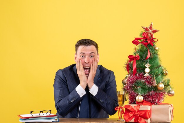 Vooraanzicht van opgetogen man handen op zijn wang zittend aan de tafel in de buurt van kerstboom en presenteert op geel