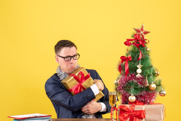 Vooraanzicht van opgewonden man die zijn geschenk stevig vasthoudt aan de tafel in de buurt van de kerstboom en geschenken op gele muur kopie ruimte