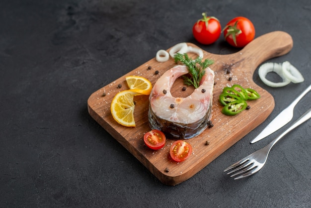 Vooraanzicht van rauwe vis en verse gehakte groenten, citroenschijfjes, kruiden op een houten bord aan de linkerkant op een zwart, verontrust oppervlak