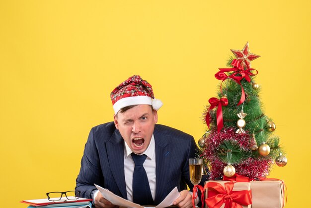 Vooraanzicht van schreeuwde man met sana zittend aan de tafel in de buurt van kerstboom en cadeautjes op geel
