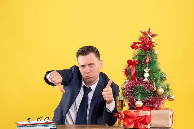 Vooraanzicht van serieuze man die duim op en neer teken zittend aan de tafel in de buurt van kerstboom en geschenken op geel