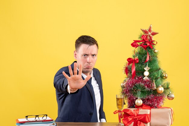 Vooraanzicht van serieuze man met stopbord zittend aan de tafel in de buurt van kerstboom en presenteert op geel