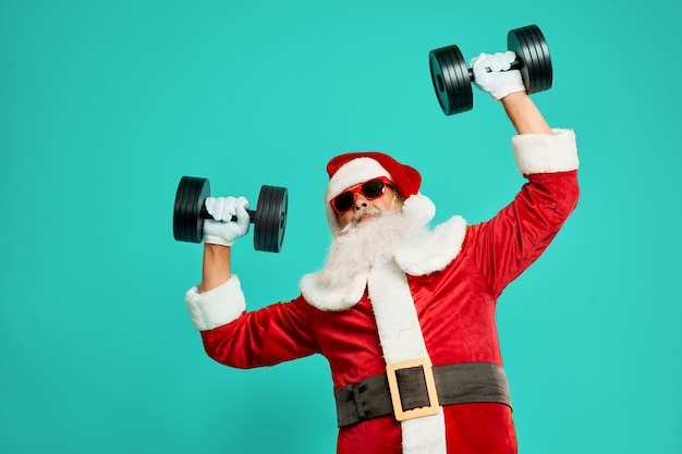 Vooraanzicht van sportieve Santa Claus-holding dumbbels. Geïsoleerde gewas van grappige senior man in kerst kostuum en zonnebril poseren