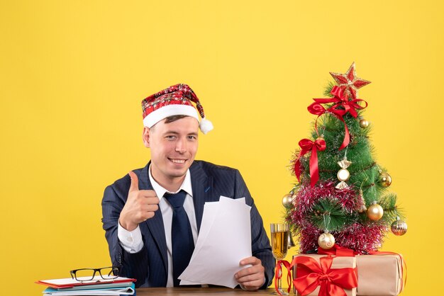 Vooraanzicht van tevreden man die duim omhoog teken zittend aan de tafel in de buurt van de kerstboom en presenteert op geel