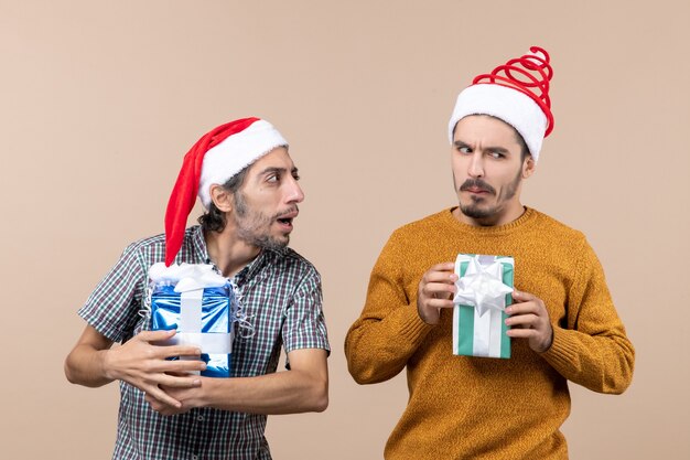 Vooraanzicht van twee verwarde jongens die kerstmutsen dragen en cadeautjes op beige geïsoleerde achtergrond houden