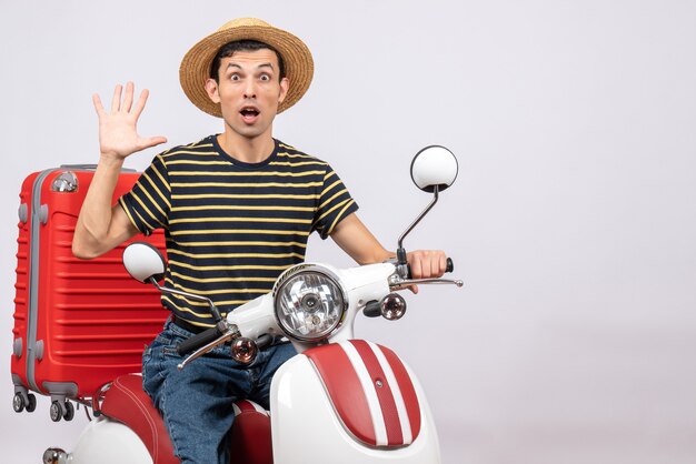 Vooraanzicht van verbaasde jonge man met strooien hoed op bromfiets zwaaiende hand