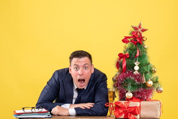 Vooraanzicht van verbaasde man die zijn mond opent aan de tafel in de buurt van de kerstboom en presenteert op gele muur