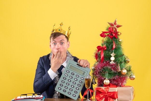 Vooraanzicht van verbaasde man met rekenmachine zittend aan de tafel in de buurt van kerstboom en presenteert op gele muur