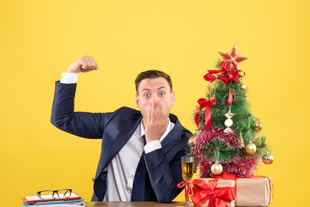 Vooraanzicht van verbaasde man met spier zittend aan de tafel in de buurt van kerstboom en presenteert op gele muur