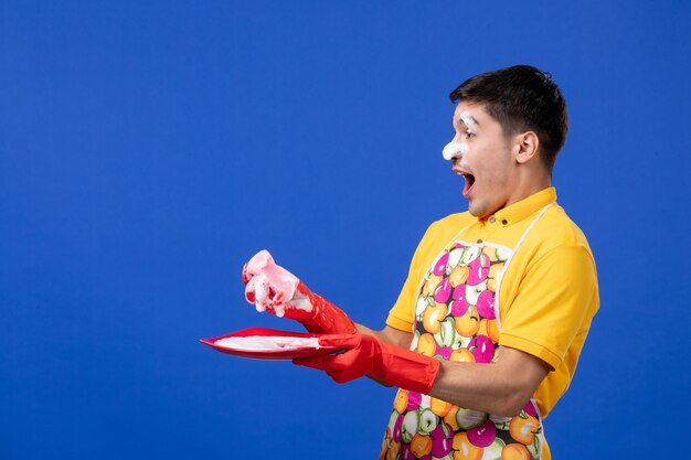 Vooraanzicht van verheugde huishoudster met schuim op zijn gezicht die plaat op blauwe muur wast