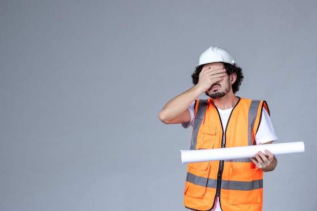 Vooraanzicht van vermoeide mannelijke constructeur in waarschuwingsvest met veiligheidshelm en leeg op grijze muur