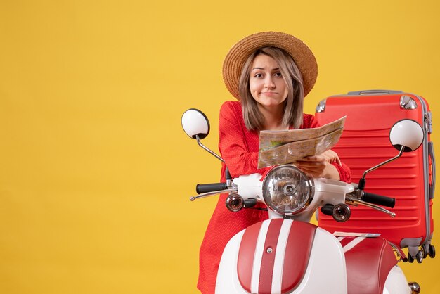 Vooraanzicht van verwarde jonge dame in rode jurk met kaart in de buurt van bromfiets