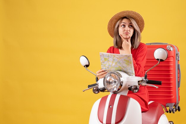 Vooraanzicht van verwarde jonge dame op bromfiets met rode koffer met kaartje