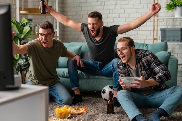 Gratis foto vooraanzicht van vrolijke mannelijke vrienden die samen naar sport op tv kijken terwijl ze snacks en bier hebben