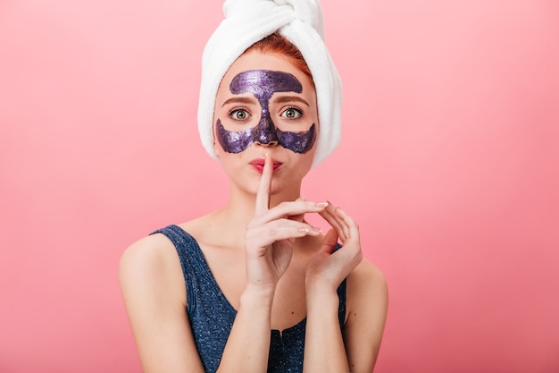 Vooraanzicht van vrouw met gezichtsmasker dat stilte teken toont. Studio shot van jonge dame in handdoek poseren op roze achtergrond tijdens huidverzorging behandeling.