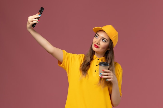 Vooraanzicht van vrouwelijke koerier in geel uniform en pet die de koffiekop van de levering houden en een foto op roze muur nemen