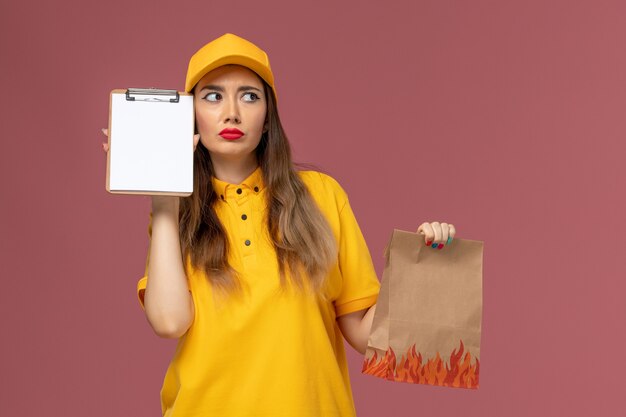 Vooraanzicht van vrouwelijke koerier in geel uniform en pet die voedselpakket en blocnote houden die aan de roze muur denken