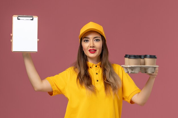 Vooraanzicht van vrouwelijke koerier in geel uniform en pet met bruine koffiekopjes en blocnote op de roze muur