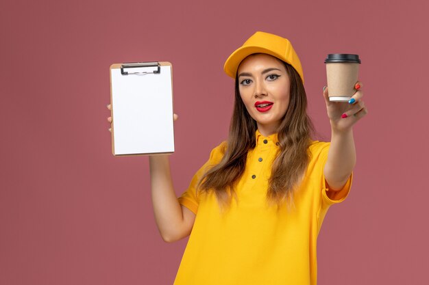 Vooraanzicht van vrouwelijke koerier in geel uniform en pet met koffiekopje en blocnote op de roze muur