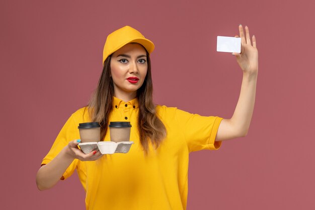 Vooraanzicht van vrouwelijke koerier in geel uniform en pet met koffiekopjes en kaart op roze muur