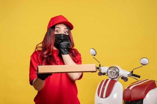 Vooraanzicht van vrouwelijke koerier in masker met de doos van het leveringsvoedsel op gele muur