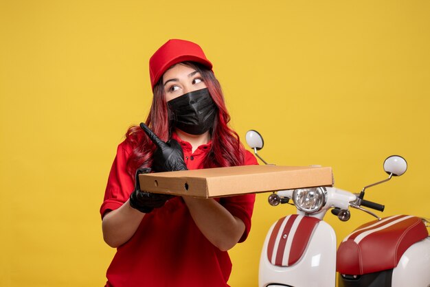 Vooraanzicht van vrouwelijke koerier in masker met de doos van het leveringsvoedsel op gele muur