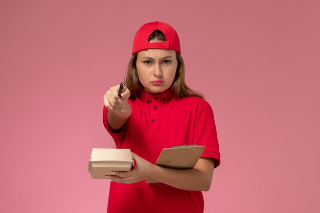 Vooraanzicht van vrouwelijke koerier in rood uniform en cape die weinig voedselpakket en blocnote houdt voor het schrijven van notities op roze muur, uniforme bezorgdienst