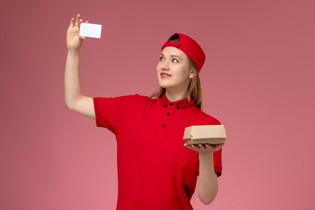 Vooraanzicht van vrouwelijke koerier in rood uniform en cape die weinig voedselpakket voor bezorging met witte plastic kaart op roze muur houdt