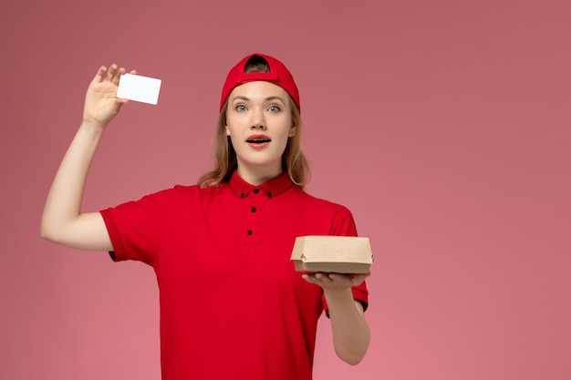 Vooraanzicht van vrouwelijke koerier in rood uniform en cape met klein voedselpakket voor bezorging met witte plastic kaart op de roze muur, service-uniforme bezorgopdracht