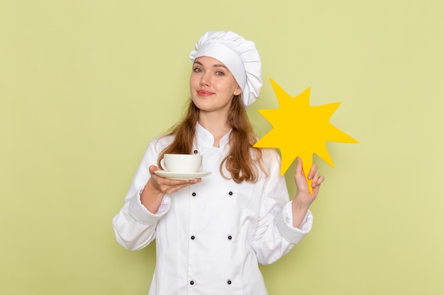 Vooraanzicht van vrouwelijke kok die wit kokkostuum draagt dat geel teken met kop van koffie op groen bureau houdt keuken keuken koken maaltijd vrouwelijke kleur