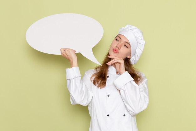 Vooraanzicht van vrouwelijke kok die wit kokkostuum draagt dat groot teken houdt en aan groene muur denkt