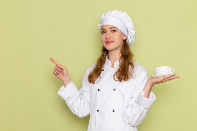 Vooraanzicht van vrouwelijke kok in wit kokkostuum die kop van koffie houden en op groen bureau keuken keuken koken maaltijd vrouwelijke kleur glimlachen