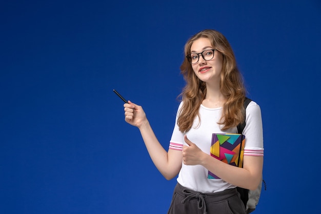 Vooraanzicht van vrouwelijke student in de witte pen en voorbeeldenboek van de overhemdsholding op het blauwe bureau