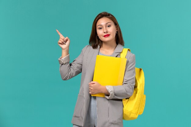 Vooraanzicht van vrouwelijke student in grijs jasje die haar gele rugzak draagt en dossiers op de lichtblauwe muur houdt