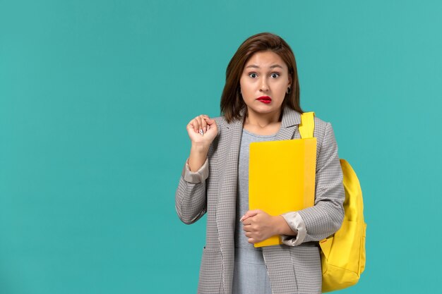 Vooraanzicht van vrouwelijke student in grijs jasje die haar gele rugzak draagt en dossiers op de lichtblauwe muur houdt