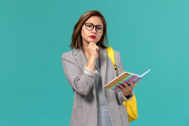 Vooraanzicht van vrouwelijke student in grijs jasje die haar gele voorbeeldenboek van de rugzakholding op lichtblauwe muur dragen