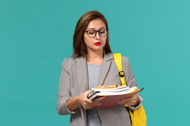 Vooraanzicht van vrouwelijke student in grijze jas gele rugzak met boeken op de blauwe muur