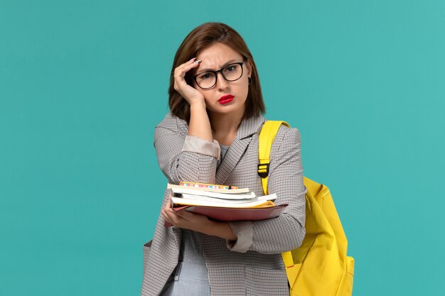Vooraanzicht van vrouwelijke student in grijze jas gele rugzak met boeken op de blauwe muur