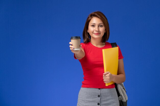 Vooraanzicht van vrouwelijke student in rood overhemd die met rugzak gele dossiers en koffie op blauwe muur houden