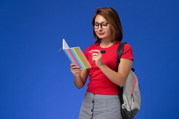 Vooraanzicht van vrouwelijke student in rood overhemd die met rugzak viltstiften houden die voorbeeldenboek op de blauwe muur lezen